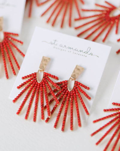 Bold coral and gold Sunburst Statement Drop Earrings with hammered gold accents are displayed on a white "st. armands designs of sarasota" card, with additional pairs softly blurred in the background.