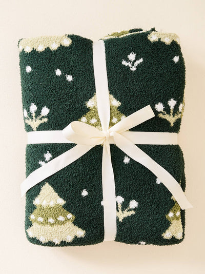 Introducing the Cozy Dreams Luxe Blanket-Christmas Trees, a dark green knitted masterpiece. Folded neatly and tied with a white ribbon, it showcases festive patterns of white snowflakes and green Christmas trees topped with snow. Experience its luxurious softness wrapped in a delightful Christmas theme.