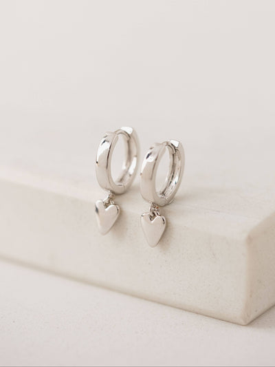 Close-up of the Everly Heart Huggie Drop Hoop Earrings in silver. These rhodium-plated hoops feature a delicate dangling heart charm, combining elegance with a playful touch.