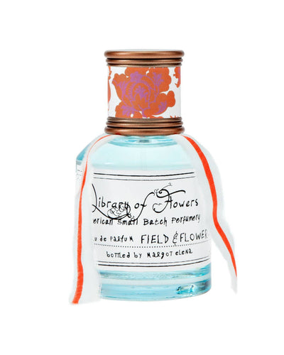 A glass bottle of Field & Flowers Eau de Parfum from the Library of Flowers collection, featuring a copper-toned cap wrapped in floral-patterned paper and a delicate fabric ribbon tied around the neck. The label displays elegant handwritten-style text.