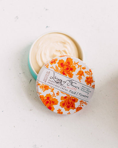 An open tin of Field & Flowers Parfum Crema, revealing the rich, smooth cream inside. The floral-patterned lid is slightly tilted, displaying the elegant packaging against a white surface.