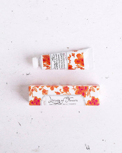 A tube of Field & Flowers Petite Treat Handcreme with orange floral packaging, placed on a white surface. The matching decorative box is positioned below the tube, displaying the "Library of Flowers" branding in elegant script.