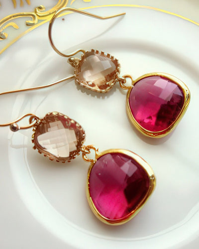 A pair of striking earrings featuring 13mm faceted fuchsia teardrop gems and 9mm champagne peach square blocks, set in 16k gold-plated bezels with 14k gold-filled earwires. Displayed on a white plate with gold detailing, these earrings exude bold elegance.