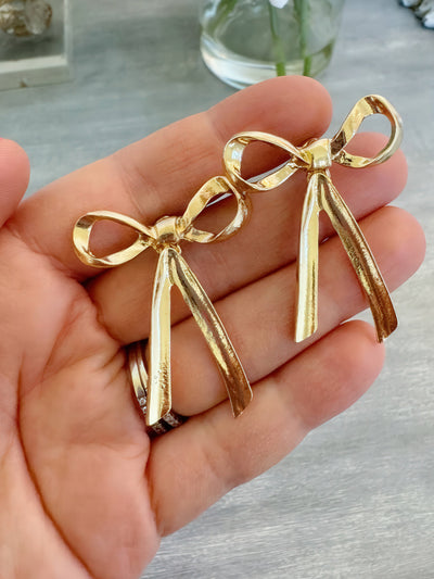 A pair of gold stud bow earrings measuring approximately 1.5 inches, with a shiny and textured finish, displayed resting on a hand. These earrings are perfect for gifting or adding a touch of elegance to everyday outfits.