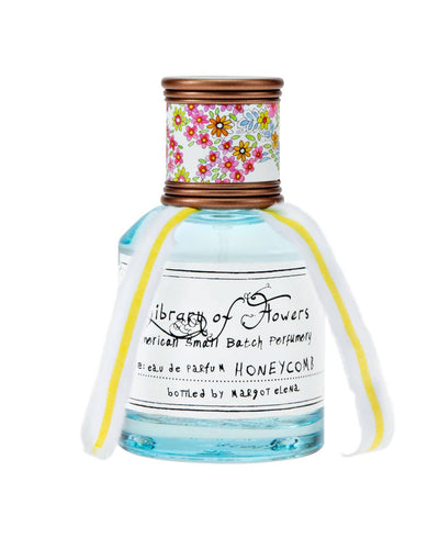 A close-up of a glass perfume bottle labeled "Library of Flowers Honeycomb Eau de Parfum," adorned with a bronze cap featuring a floral pattern and a white-and-yellow ribbon tied around the neck.