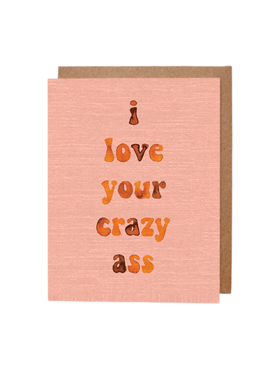 A peach-toned greeting card with subtle textured detailing featuring the phrase "i love your crazy ass" written in a playful, orange and brown gradient font. The card is accompanied by a brown kraft envelope in the background.