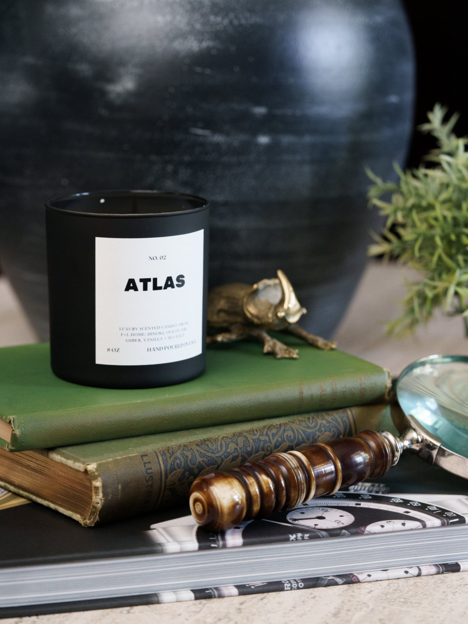 A black No. 02 Atlas 8oz Soy Luxury Candle from F+L Home rests on two green hardcover books. Nearby, a magnifying glass, a wooden-handled tool, and a small metallic dinosaur figurine are artfully placed. In the background, a portion of a large dark vase and some greenery enhance the ambiance of bespoke scents and elegance.