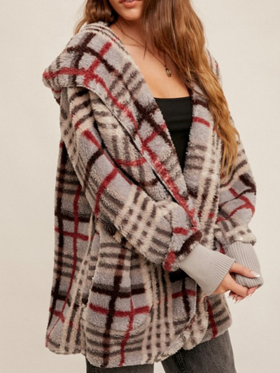 A person is enveloped in a Teddy Open Fur Jacket - Grey Plaid, showcasing its cozy checkered design with hues of gray, black, and red. Their long hair falls over a black top paired with dark pants, highlighting the warmth of the jacket.