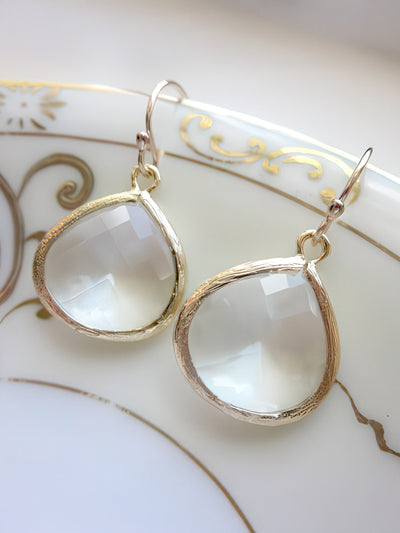 A pair of large crystal teardrop earrings featuring 15x18.5mm faceted gems set in gold-plated bezels with 14k gold-filled earwires. Displayed on a white ceramic dish with gold detailing, these earrings exude timeless elegance.