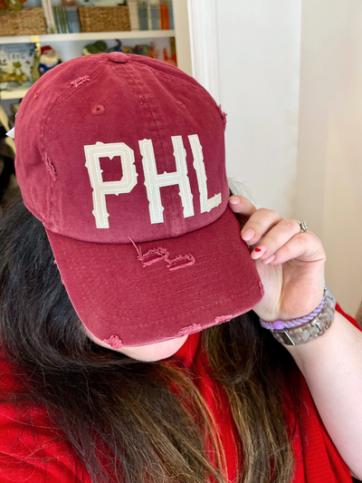 Sporting a PHL Distressed Vintage-Inspired Baseball Hat in maroon with white "PHL" letters, a person holds the brim, showcasing their ring and watch. The hat complements their red top as books and other items blur in the background, highlighting their Philly pride.