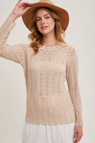 Woman wearing a beige sheer knit long-sleeve top with intricate detailing, styled with a white skirt and wide-brim hat for a soft, bohemian-inspired look.