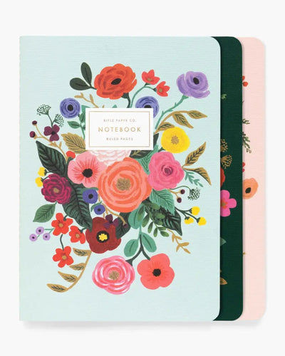 Set of three Rifle Paper Co. stitched notebooks featuring vibrant floral illustrations on soft pastel and dark-toned covers, with gold foil accents and ruled pages for journaling and note-taking.