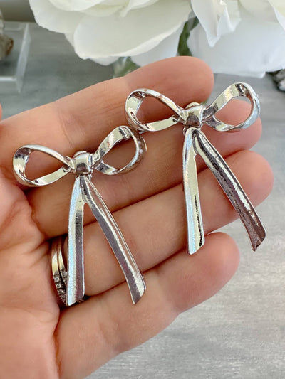 A pair of silver stud bow earrings measuring approximately 1.5 inches, with a shiny and polished finish, displayed resting on a hand. These earrings are perfect for gifting or as a stylish addition to everyday outfits.