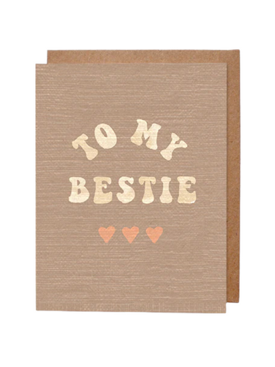 A beige greeting card with a subtle textured design featuring the phrase "TO MY BESTIE" written in gold letters with a playful font. Below the text are three small pink hearts in a row. A brown kraft envelope is partially visible in the background.