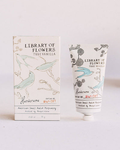 A tube of True Vanilla Handcreme standing next to its matching box, both featuring an elegant illustration of birds and branches in soft blue and brown hues. The packaging highlights the Library of Flowers branding and small-batch perfumery details.