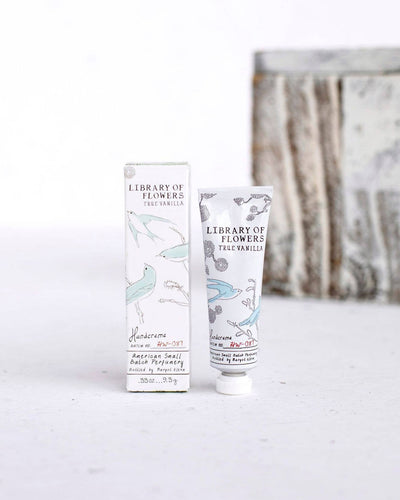 A tube of Library of Flowers True Vanilla Petite Treat Handcreme standing next to its matching box. The packaging features delicate illustrations of bluebirds and botanical elements against a soft cream background, highlighting the elegant and artistic design of this small-batch perfumery product.