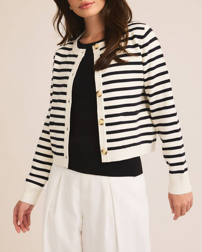 Woman wearing a white and navy striped button-down cardigan with tortoiseshell buttons and a round neckline, styled with a black top and white trousers for a classic, nautical-inspired look