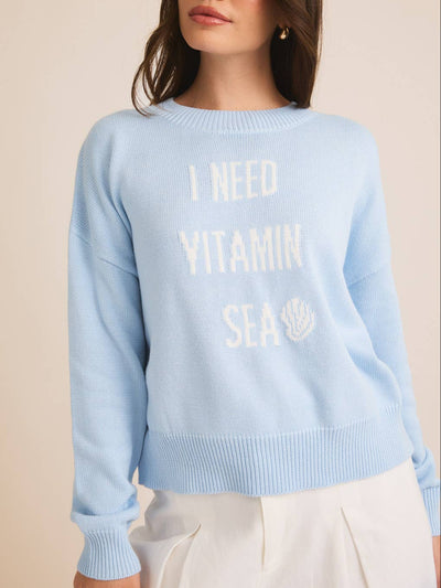 Wearing an "I NEED VITAMIN SEA" Sweater in baby blue with a seashell graphic, a woman sets off her long dark hair with a white skirt against a neutral backdrop, ideal for beach lovers.